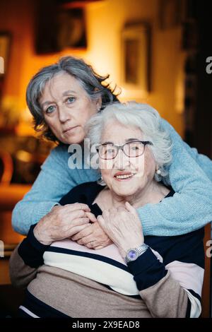 Porträt einer älteren Frau, die lächelt, während ihre Tochter sie von hinten umarmt. Beide sehen zufrieden aus und zeigen ihre starke Verbundenheit in einem gemütlichen Setting Stockfoto