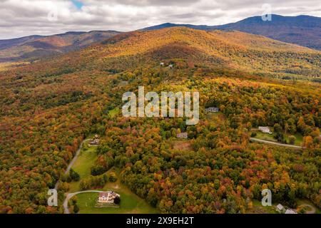 Der Herbstwald schmückt das Dorf unter Wolken vor den Bergen. Drohnenfoto. Vermont, USA Stockfoto