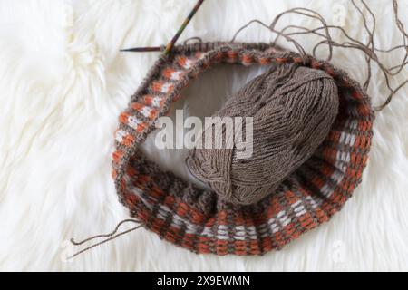 Flachbild-Fotografie von Fair Isle-Strickarbeiten mit braunem Alpaka-Garn auf bunten Holznadeln auf weißem Lammfellfell Stockfoto