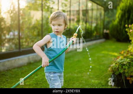 Süßer kleiner Junge, der am Sommertag im Garten Blumenbeete gießt. Kind, das Gartenschlauch zum Gießen von Gemüse verwendet. Kind hilft bei alltäglichen Aufgaben. Momm Stockfoto