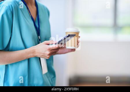 Medizinischer Fachmann in Scrubs mit Smartphone während der Kaffeehaltung im Krankenhaus Stockfoto