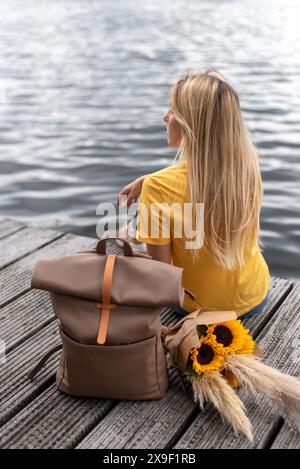 Eine junge Frau sitzt auf einem Pier am See neben dem Rucksack eines Reisenden und einem Blumenstrauß. Stockfoto