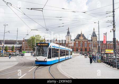 Eine blau-weiße Straßenbahn, die am 28. Mai 2024 abgebildet wurde und eine Haltestelle außerhalb des Amsterdamer Centraal, Niederlande, ablegt, während Touristen und Pendler auf den Wegen laufen. Stockfoto