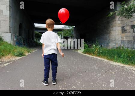 Rückansicht kleiner niedlicher Junge, der einen roten Ballon hält, steht vor einem dunklen Betontunnel auf einer leeren Stadtstraße. Allein verlor tapferes Kind Stockfoto