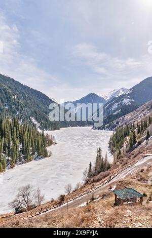 Almaty, Kasachstan - 19. März 2024: Fußgängerweg aus Holz in der Nähe des malerischen eisbedeckten Kolsai-Sees, umgeben von grünen Hügeln. Feder Stockfoto