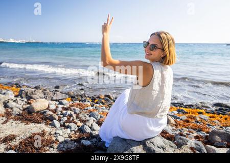 Eine Frau sitzt auf einem Felsen am Meer, trägt eine Sonnenbrille und macht das Friedenszeichen Stockfoto