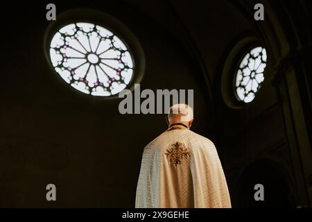Rückansicht eines nicht wiedererkennbaren älteren katholischen Priesters, der in der Kirche mit runden Fenstern steht Stockfoto