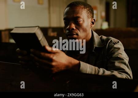 Junger afroamerikanischer Mann, der auf einer Bank in der katholischen Kirche sitzt und das Bibelbuch in der Hand hält, mittlere Nahaufnahme Stockfoto