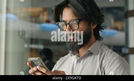 Fokussierter seriöser indischer Mann bärtiger Geschäftsmann mit Brille männlicher Kunde, der Handy benutzt, um E-Mails zu lesen, in sozialen Netzwerken zu surfen und Medien zu lesen Stockfoto