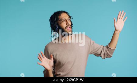 Unbeschwerter Mann aus dem Nahen Osten, der energische Musik hört, vom Rhythmus schwärmt und Freizeit genießt. Euphorische BIPOC-Person, die Kopfhörer trägt, Tanzbewegungen macht, isoliert über Studiohintergrund Stockfoto