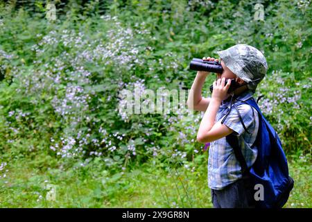 Kind von 7-8 Jahren, Junge in Panama Hut, kariertes Hemd mit Walkie-Talkie in der Hand steht im Wald, schaut sorgfältig durch das Fernglas, Konzept des Wanderens Stockfoto