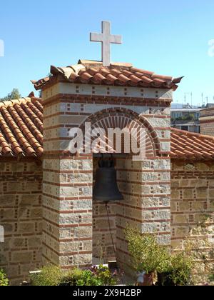 Kleine Kirche mit Glocke und Kreuz darüber umgeben von Pflanzen unter klarem Himmel, antike Gebäude mit Säulen und Bäumen auf der Akropolis in Athen Stockfoto