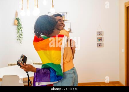 Schwules lateinisches, junges Paar, das in LGBT-Flagge gehüllt ist, während es Fotos in der Wohnung macht Stockfoto