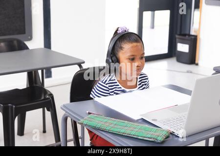 In der Schule, eine junge birassische Schülerin, die Kopfhörer trägt und sich im Klassenzimmer auf einen Laptop konzentriert Stockfoto