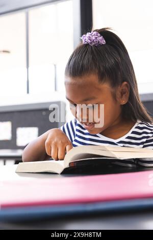 In der Schule, ein junges birassisches Mädchen, das sich darauf konzentriert, ein Buch im Klassenzimmer zu lesen Stockfoto