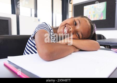 In der Schule legt sich ein junges birassisches Mädchen mit einem hellen Lächeln im Klassenzimmer auf die Arme Stockfoto