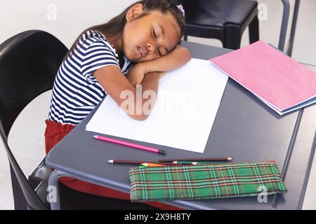 In der Schule lag ein junges birassisches Mädchen mit einem gestreiften Hemd im Klassenzimmer mit dem Kopf auf dem Schreibtisch Stockfoto