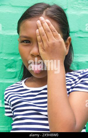 In der Schule, draußen, ein birassisches junges Mädchen, das ein Auge bedeckt und die Zunge herausstreckt Stockfoto