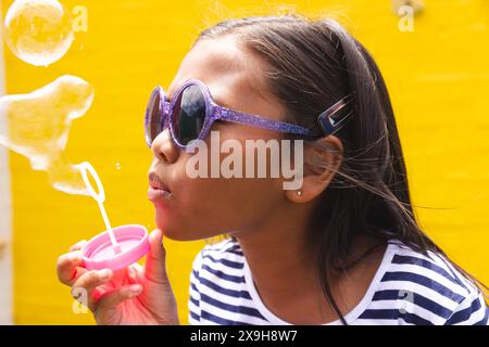 In der Schule bläst ein junges birassisches Mädchen, das eine Sonnenbrille trägt, draußen Blasen. Sie hat dunkles Haar, hellbraune Haut und trägt ein gestreiftes Oberteil. Stockfoto