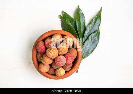 Ein Haufen frischer reifer Litschi-Früchte auf einem Tontopf mit grünen Blättern auf weißem hölzernem Hintergrund. Blick von oben auf die gesunde tropische Frucht Litchi chinensis. Stockfoto