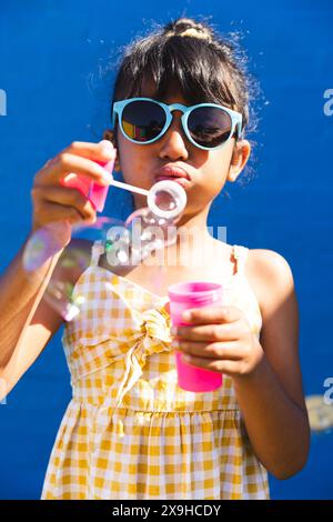 Ein birassisches Mädchen mit Sonnenbrille und Sommerkleid bläst draußen Blasen Stockfoto