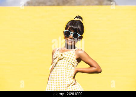 Ein birassisches Mädchen, das eine Sonnenbrille und ein Sommerkleid trägt, posiert selbstbewusst im Freien Stockfoto