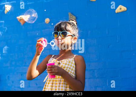 Ein birassisches Mädchen, das eine Sonnenbrille und ein Sommerkleid trägt, bläst gern draußen Blasen, mit Kopierraum Stockfoto