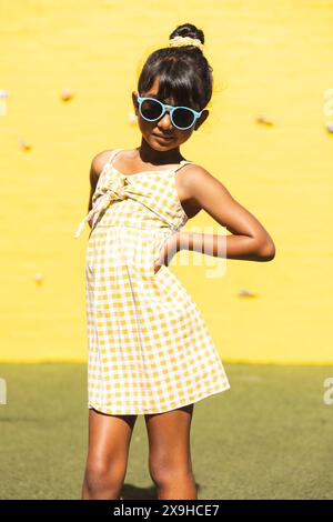 Ein birassisches Mädchen, das eine Sonnenbrille und ein Sommerkleid trägt, posiert selbstbewusst im Freien Stockfoto