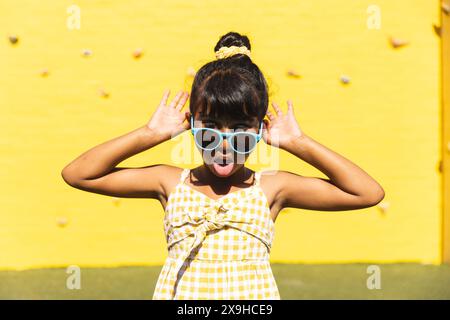 Ein birassisches Mädchen, das eine Sonnenbrille und ein Sommerkleid trägt, macht draußen ein verspieltes Gesicht Stockfoto