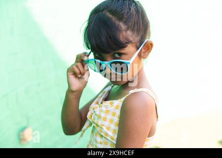 Ein birassisches Mädchen, das eine Sonnenbrille und ein Sommerkleid trägt, posiert spielerisch im Freien Stockfoto