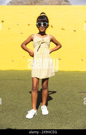 Ein birassisches Mädchen, das eine Sonnenbrille und ein Sommerkleid trägt, posiert selbstbewusst im Freien Stockfoto