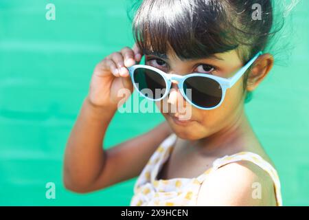 Ein birassisches Mädchen, das eine Sonnenbrille und ein Sommerkleid trägt, posiert spielerisch im Freien Stockfoto