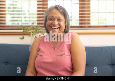 Zu Hause lächelt eine zweirassige Seniorin während eines Videogesprächs auf der blauen Couch Stockfoto