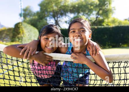 Im Freien lächeln zwei junge Schwestern und lehnen sich an sonnigen Tagen auf das Tennisnetz Stockfoto