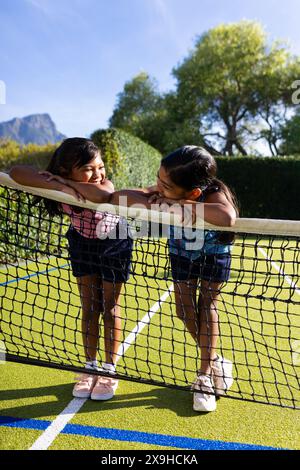 Draußen lehnen sich zwei junge Schwestern auf ein Tennisnetz und lächeln einander an Stockfoto