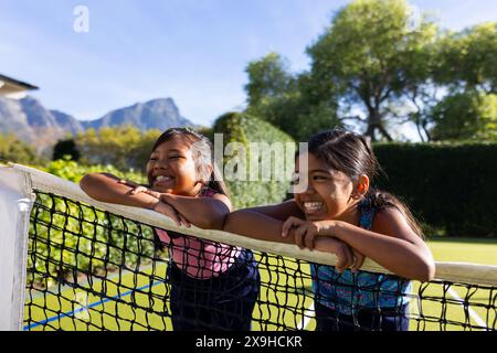 Im Freien, zwei birassische junge Schwestern, die sich auf das Tennisnetz stützen, lächeln und Spaß haben Stockfoto