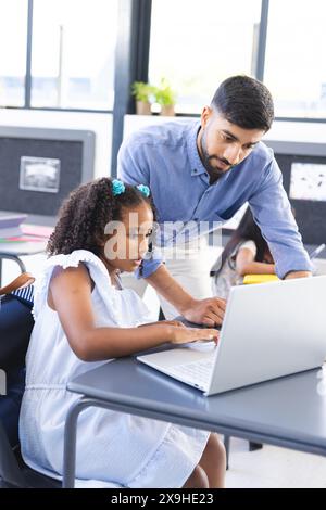 Asiatischer männlicher Lehrer hilft jungen, birassischen Schülerinnen mit Laptop im Klassenzimmer Stockfoto