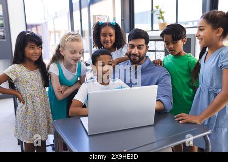 In der Schule versammeln sich junge asiatische Lehrer und verschiedene Schüler um einen Laptop im Klassenzimmer Stockfoto