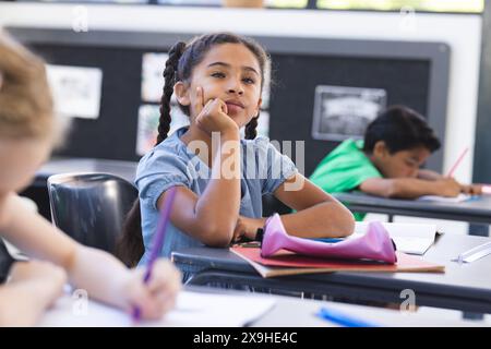 Ein birassisches Mädchen mit geflochtenen Haaren sieht nachdenklich aus in einer Schulklasse. Sie ist von Gleichaltrigen umgeben, die während eines Unterrichts eine Schule vorschlagen, betonen Stockfoto