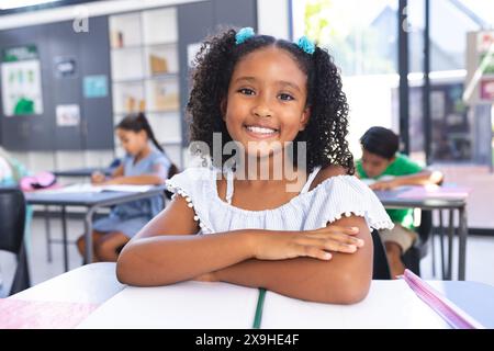 In der Schule lächelt ein junges birassisches Mädchen mit lockigen schwarzen Haaren an ihrem Schreibtisch Stockfoto