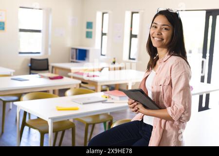 Junge birassische Frau steht selbstbewusst in einem Klassenzimmer in der High School. Sie ist eine Lehrerin, die ein Tablet hält und bereit ist, Schüler mit Technologie in e zu interagieren Stockfoto