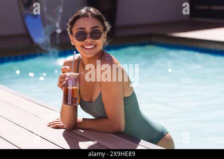 Eine zweirassige junge Frau im Freien, lehnt sich am Pool und hält ein Glas Eistee Stockfoto