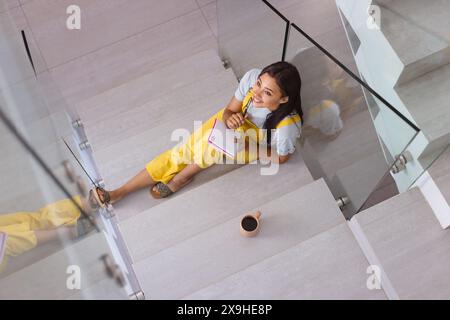 Eine zweirassige junge Frau, die bunte Notizbücher zu Hause hält und einen gelben Jumpsuit trägt Stockfoto