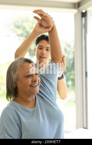 Ein junger birassischer Physiotherapeut hilft einer älteren Patientin, zu Hause Sport zu treiben. Heller, luftiger Raum mit großen Fenstern, die draußen Grün zeigen, c Stockfoto