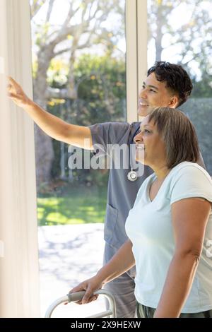 Zu Hause, männliche Krankenschwester und ältere Patientin mit Gehhilfe. Sie sind in einem hellen Zimmer mit großen Fenstern mit Blick auf einen üppigen Garten, unaltere Stockfoto