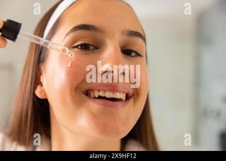 Zu Hause, eine junge kaukasische Frau, die ihr Hautpflegeserum auf das Gesicht aufträgt, lächelnd. Sie ist in einem hellen, modernen Badezimmer mit weißem Hintergrund und genießt ein Zuhause Stockfoto