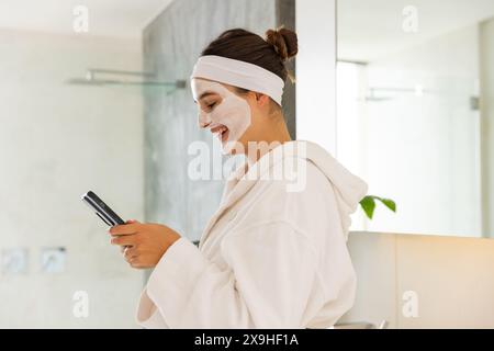 Zu Hause eine junge Kaukasierin mit einer Gesichtsmaske im Bademantel mit Smartphone. Sie ist in einem modernen, hellen Badezimmer mit einem großen Spiegel und Mini Stockfoto