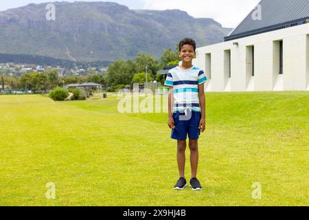 Biracial Boy steht lächelnd auf einem sonnigen Grasfeld, Berge im Hintergrund, mit Kopierraum Stockfoto