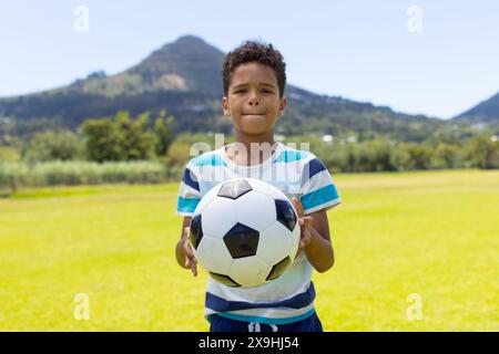 Biracial Boy hält einen Fußball, steht auf einem sonnigen Feld mit Bergen dahinter Stockfoto