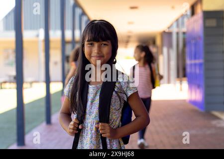 Ein birassisches Mädchen mit langen dunklen Haaren lächelt in der Schule und hält ihre Rucksackgurte Stockfoto